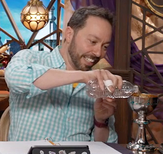 Sam pouring water from a plastic disposable water bottle into a silver chalice. The chalice is reflective, with a wide ridge halfway up its stem. The inside is a dull golden bronze. Sam’s face is set in concentration.