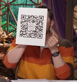 A piece of printer paper with a QR code printed on it, tilted sideways. It is attached to the base of a vessel that Sam is drinking from, although the paper hides the vessel itself. The Critical Role logo is in the center of the QR code. The QR code leads to samsamericangirldollshoes.com.