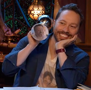 Sam holding a silver chalice to reveal an image of Brennan on the bottom. Brennan is looking deviously forward. A number of injuries or substances on his face are difficult to make out given the image quality. Sam smiles mischievously, his chin resting on the upturned palm of his hand.