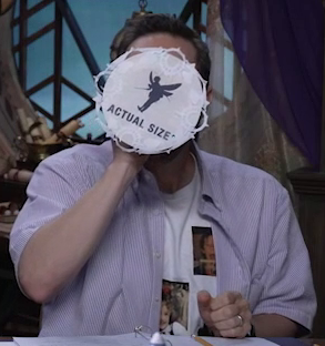 A circular piece of paper with a silhouette of Thimble on the bottom. Under the silhouette is the text “ACTUAL SIZE”. The paper is attached to the base of a vessel that Sam is drinking from, although the paper hides the vessel itself. Circular decorative patterns are cut out and pasted around the edges of the paper.