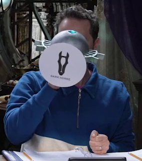 Sam drinking from a large ornate silver chalice with the Dark Horse Wine logo, a black horse head, pasted on the bottom, the text “DARK HORSE” below it. The chalice has spokes emerging from the curve of its cup reminiscent of Candescent Creed sun ray designs. A turquoise gem is set in the side, matching the gem in Wicander’s circlet. Sam tips the chalice fully horizontally into his mouth.