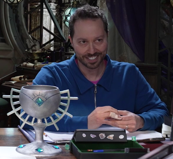 Sam sitting at the table behind a large ornate silver chalice. The chalice has spokes emerging from the curve of its cup reminiscent of Candescent Creed sun ray designs. A turquoise gem is set in the side with more sun ray designs drawn in black around it, exactly matching Wicander’s circlet. Four more turquoise gems are set around the wide base of the chalice. Sam looks forward, mischief on his face. A splint is on his right ring finger.