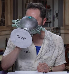 Sam drinking from a large ornate silver chalice with the next “POOP” written in black marker on white paper on the bottom. The chalice has spokes emerging from the curve of its cup reminiscent of Candescent Creed sun ray designs. A turquoise gem is set in the side, matching the gem in Wicander’s circlet. Sam wears a thin white button-up shirt opened to reveal the top of his chest and a royal blue shirt underneath.