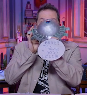 Sam drinking from a large ornate silver chalice with the text “BRRR!! THIS OPENING IS SOOO COLD!!!” hand-written on white paper pasted to the bottom. The chalice has spokes emerging from the curve of its cup reminiscent of Candescent Creed sun ray designs. A turquoise gem is set in the side, matching the gem in Wicander’s circlet. Sam wears a tan snap-up jacket over a black T-shirt with a white graphic across the front, not fully visible under the snap-up.