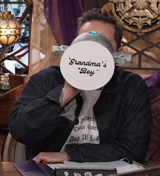 Sam drinking from a large ornate silver chalice with the text “Grandma’s Boy” printed on white paper pasted to the bottom. The text is in a cursive font and has a few sparkles surrounding it. The chalice has spokes emerging from the curve of its cup reminiscent of Candescent Creed sun ray designs. A turquoise gem is set in the side, matching the gem in Wicander’s circlet. Sam tips the chalice horizontally into his mouth.