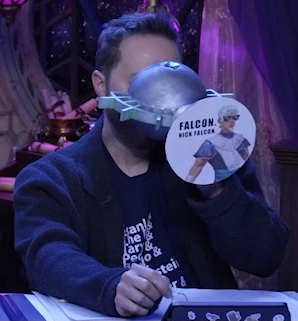 Sam drinking from a large ornate silver chalice with an image of Wicander wearing sunglasses and the text “FALCON. NICK FALCON” printed on white paper pasted to the bottom. The chalice has spokes emerging from the curve of its cup reminiscent of Candescent Creed sun ray designs. A turquoise gem is set in the side, matching the gem in Wicander’s circlet. Sam tips the chalice horizontally into his mouth.