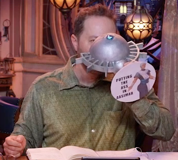 Sam drinking from a large ornate silver chalice with an image of Wicander with an enlarged butt and the text “PUTTING THE ASS IN AASIMAR” printed on white paper pasted to the bottom. The chalice has spokes emerging from the curve of its cup reminiscent of Candescent Creed sun ray designs. A turquoise gem is set in the side, matching the gem in Wicander’s circlet. Sam tips the chalice horizontally into his mouth.