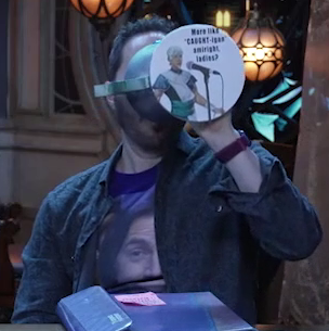 Sam drinking from a large ornate silver chalice with an image of Wicander at a stand-up microphone and the text “More like ‘CAUGHT’-igan amiright ladies?” printed on white paper pasted to the bottom. The chalice has spokes emerging from the curve of its cup reminiscent of Candescent Creed sun ray designs. A turquoise gem is set in the side, matching the gem in Wicander’s circlet. Sam tips the chalice horizontally into his mouth.