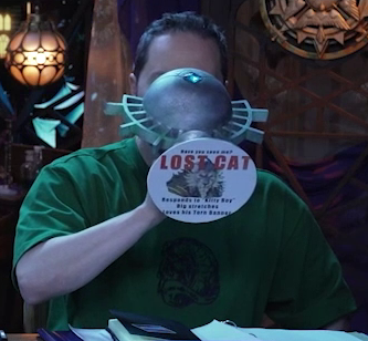 Sam drinking from a large ornate silver chalice with an image of Teor and the text “Have you seen me? LOST CAT. Responds to ‘Kitty Boy’. Big stretches” and then a final line that isn’t readable. This is printed on white paper pasted to the bottom of the chalice. The chalice has spokes emerging from the curve of its cup reminiscent of Candescent Creed sun ray designs. A turquoise gem is set in the side, matching the gem in Wicander’s circlet. Sam tips the chalice horizontally into his mouth.