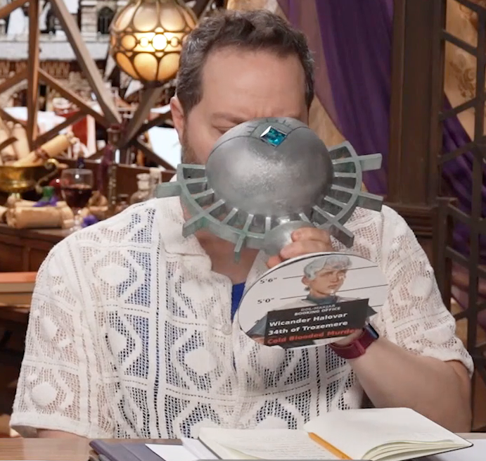 Sam drinking from a large ornate silver chalice with a mug shot of Wicander behind a placard reading “DOL-MAKJAR BOOKING OFFICE. Wicander Halovar. 34th of Trozemere. Cold Blooded Murder”. The words “Cold Blooded Murder” are in red, the rest of the text is in white. The chalice has spokes emerging from the curve of its cup reminiscent of Candescent Creed sun ray designs. A turquoise gem is set in the side, matching the gem in Wicander’s circlet. Sam tips the chalice into his mouth.