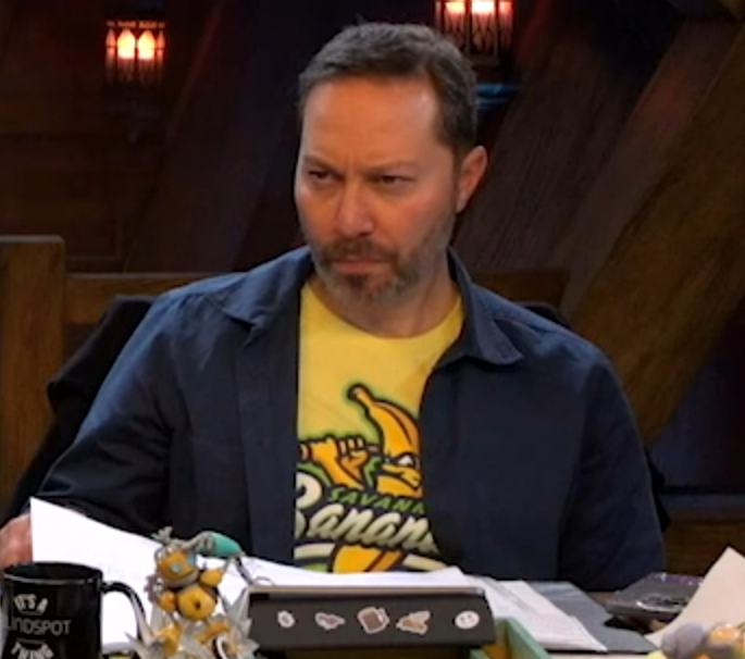 Sam wearing a yellow T-shirt mostly covered dark navy collared long-sleeve button-up. The shirt depicts an intense-looking anthropomorphic banana swinging a baseball bat. The visible text seems to read “SAVAGE Banana”. Sam is squinting, looking sternly in front of him.