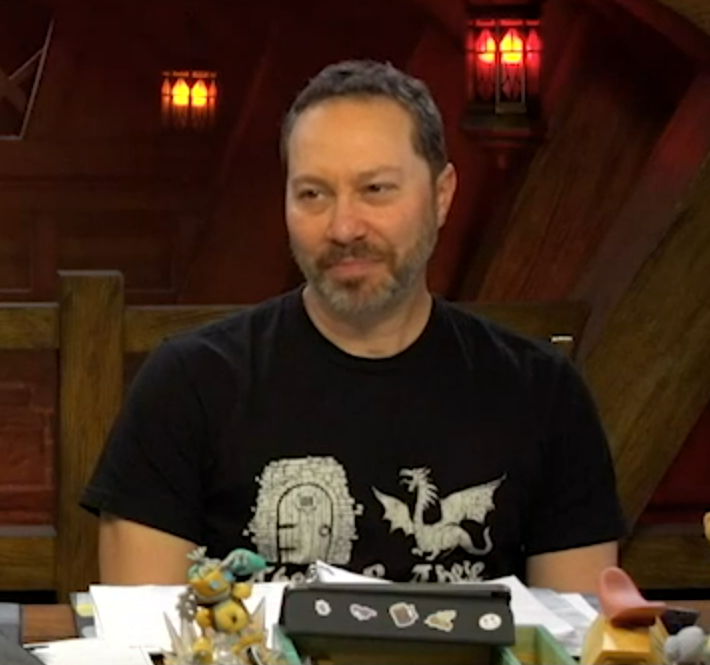 Sam wearing a black T-shirt depicting a white medieval-European-style door and a dragon to its right. The dragon’s mouth is open and its wings are spread. Some text is below, but is not readable due to being covered by objects on the table. Sam’s head is tilted slightly to the side looking forward.