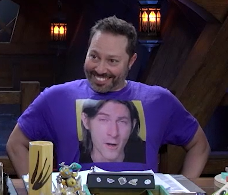 Sam wearing a purple T-shirt with a large square image of Matt centered on the chest. The image is a close up of Matt’s face, his eyes half closed and his mouth slightly open, looking goofy. Sam is grinning and eagerly displaying the shirt.