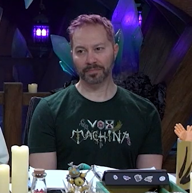 Sam wearing a forest green T-shirt that “VOX MACHINA”. The letters are stylized, where for example the “O” is a d20, the “X” is an hourglass, the “M” is a bow and arrow, and the final “A” is an axe.