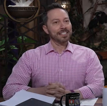 Sam wearing a business casual pink and white striped button-up shirt with the top button undone. He sits with his hands clasped on the table in front of him, paused mid speech.