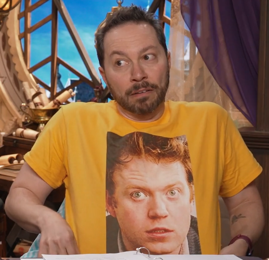 Sam sitting at the table, wearing a yellow T-shirt with an image of a wild-haired, wide-eyed Brennan printed on the front. Brennan wears a striped jacket. The image of Brennan is from the video “Tide CEO: Soap Isn’t Medicine”. Sam stretches out the shirt for visibilty, looking to his right and making a goofy frightened expression.