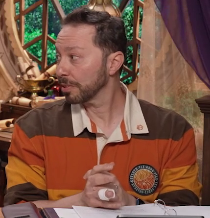 Sam sitting at the table, wearing a brown, orange, and yellow rugby shirt. A circular patch on the left breast has the names of the members of Vox Machina around its outside, and the word “VOX MACHINA” in a bubble font in the center. Sam looks to his right, speaking, hands clasped in front of him. A splint is visible on his right ring finger.