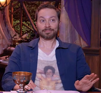 Sam sitting at the table, wearing a white T-shirt with an image of a shirtless Prince from the album Controversy. He has his hands behind his head and is standing against the tiled wall of a shower. He has voluminous black hair and a moustache. Over the T-shirt Sam has on a deep blue open button-up shirt. Sam leans back, his eyes wide in mock surprise, left hand out as if to steady himself.