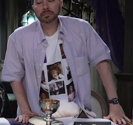 Sam standing up at the table, wearing a blue and white striped button-up shirt open to reveal a T-shirt with childhood images of the cast on it. Visible in the center is a young Ashley with curly hair and what appear to be red flowers in it. A young Laura also seems to be present in another image. Below Ashley is a black and white image of a child, and to the side of it is what seems to be a yearbook photo of a smiling child.