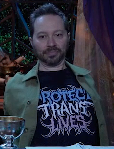 Sam sitting at the table, wearing a black T-shirt reading “PROTECT TRANS LIVES” in a heavy metal font. The words are blue, white, and pink respectively. Over the shirt sam wears a thick green button-up. Sam leans back, stretching, looking slightly worried.