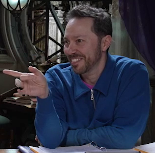 Sam sitting at the table, wearing a blue collared quarter-zip long-sleeved shirt. Underneath the shirt is something pink, only the tip of the collar visible. Sam leans forward, his elbows on the table, casually pointing to the side with his right hand. A splint is on his right ring finger.