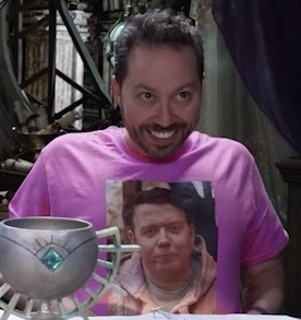 Sam sitting at the table, wearing a pink T-shirt with an image of Brennan on the front. The image is from the Dropout American Girl Doll shoes sketch. Brennan wears a knit poncho in warm earth tones and looks forward with his lips pursed. Sam straightens the shirt for improved visibility and stares with his head tilted forward, a crazed Tim Curry-like smile on his face.