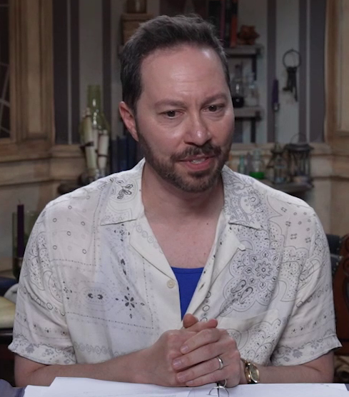 Sam sitting at the table, wearing a thin white button-up shirt opened to reveal the top of his chest and a royal blue shirt underneath. Sam sits with his hands clasped in front of him, looking down sadly and thoughtfully, in the middle of speaking. The button-up has paisley line art sparsely spread across it.