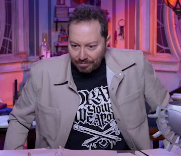 Sam sitting at the table, wearing a tan snap-up jacket over a black T-shirt reading “DRAW YOUR WEAPONS”. Under the text is art of a crossed pencil and sword, dripping with what seems to be ink. The jacket has two breast pockets, one on each side. Sam leans forward, his shoulders hunched, as if adjusting his position in his seat.