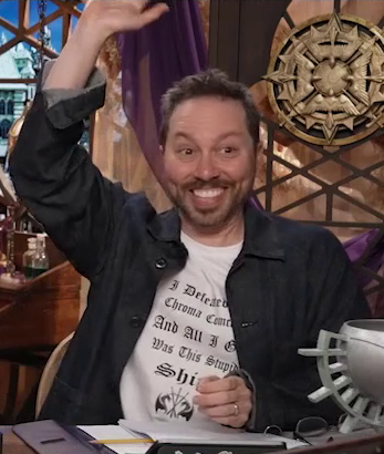 Sam sitting at the table, wearing a black button-up shirt over a white T-shirt. On the T-shirt, in black gothic font, is the text “I Defeated the Chroma Conclave And All I Got Was This Stupid Shirt”. Underneath the text is the image of two dragons facing each other with three crossed swords between them. Sam smiles, raising his right hand as if volunteering an answer in class.