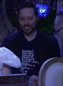 Sam sitting a bit back from the table, wearing a dark blue T-shirt with a list of his characters on it. It reads “Scanlan & The Rat & Taryon & Peddy & Benicio & Nott & Bundlestein & Slaughter & FCG & Loquatius & Pango &”. Lines below this are out of view.