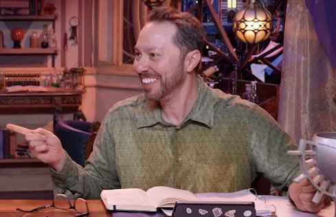 Sam sitting at the table, wearing a green long-sleeved button-up shirt with a faint diamond pattern on it. He smiles and points to his right, looking at something off-screen.