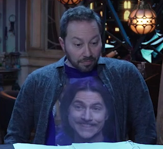 Sam sitting at the table, wearing a blue T-shirt with an image of Matt on the front. Matt grins goofily, his upper teeth biting his bottom lip in an expression resembling a beaver. Sam leans back, straightening any folds in the shirt.