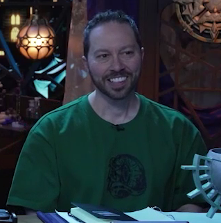 Sam sitting at the table, wearing a green T-shirt with a line-art design on it of one of Uk’otoa’s Cloven Crystals, which resembles an eyeball with a vertical slit. Sam leans forward listening, smiling slightly.