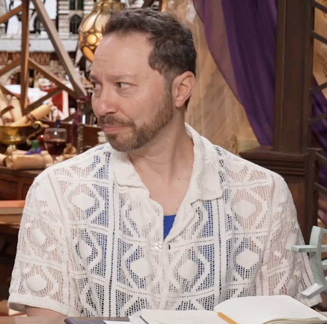Sam sitting at the table, wearing a white mesh short-sleeve button-up over a royal blue tanktop. A pattern of diamonds inlaid in squares is worked into the shirt, through which is visible Sam’s tanktop and arms. Sam looks sadly to his right, making eye contact with Whitney off-screen.