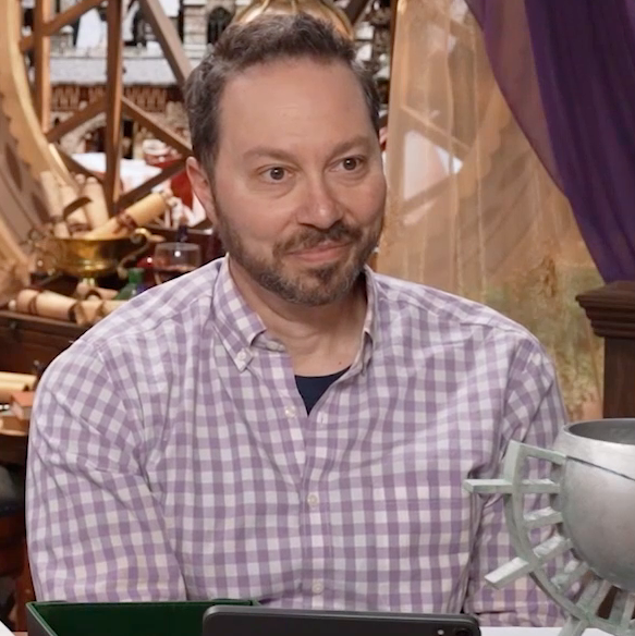 Sam sitting at the table, wearing a light purple and white checkered long-sleeve button-up with the top two buttons undone. He sits calmly, listening to someone speak, his lips pursed and eyes looking tired.