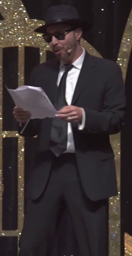 Sam standing on a stage, wearing a black suit, black necktie, black fedora, sunglasses, and a white collared shirt. He is speaking into a stage microphone attached to his cheek and reads from two stapled sheets of paper in his hands. The stage backdrop has gold glittery arches in front of a sparkly black curtain.