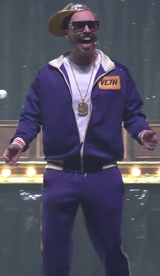 Sam standing on a stage, wearing a purple track suit with a breast patch reading “VETH”. The suit is unzipped to reveal a white T-shirt and a necklace bearing a large golden dollar symbol at its end. He wears a golden baseball cap and sunglasses, and screams into a stage microphone attached to his cheek. In the background is a DM screen set up on a table lit with a row of yellow bulbs.