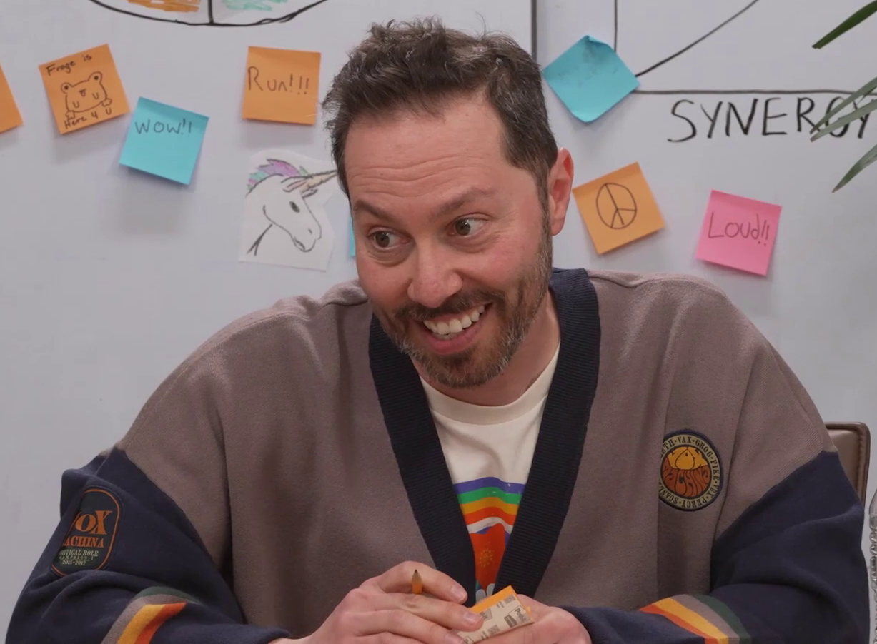 Sam wearing a roomy Vox Machina sweater over a white T-shirt bearing a rainbow and red star. He is smiling kindly and looking slightly down. The sweater is light gray with a dark blue collar and sleeves, and bands of red, yellow, gray, and green near the wrists. A patch on the right arm reads “VOX MACHINA. CRITICAL ROLL. CAMPAIGN 1. 2015–2017”. A patch on the left breast has the names of the Vox Machina members in a circle around the text “VOX MACHINA” in a bubble font. Behind him is a whiteboard with graphs, a drawing of a unicorn,  and a number of sticky notes bearing text like “Run!!!”, “Loud!!”, and “Gotta be GAY To be SLAY”.