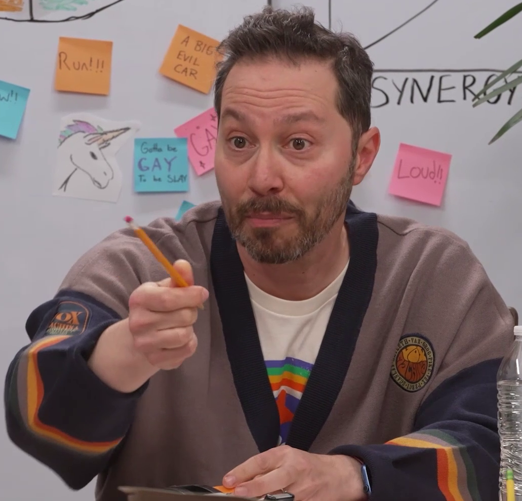 Sam wearing a roomy Vox Machina sweater over a white T-shirt bearing a rainbow and red star. He has his mouth closed in thought as he points a pencil to call on someone off-screen. The sweater is light gray with a dark blue collar and sleeves, and bands of red, yellow, gray, and green near the wrists. A patch on the right arm reads “VOX MACHINA. CRITICAL ROLL. CAMPAIGN 1. 2015–2017”. A patch on the left breast has the names of the Vox Machina members in a circle around the text “VOX MACHINA” in a bubble font. Behind him is a whiteboard with graphs, a drawing of a unicorn, and a number of sticky notes bearing text like “Run!!!”, “Loud!!”, and “Gotta be GAY To be SLAY”.