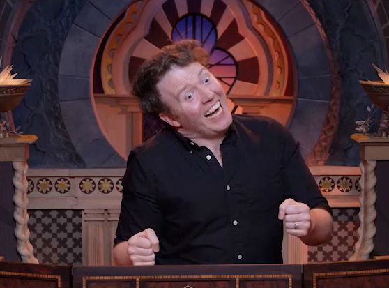 Brennan playing as Termina, making an unhinged face. He tilts his head to the side unnaturally, his eyes wide and mouth in a toothy rictus grin. He mimicks holding someone in front of him in his two clenched fists.