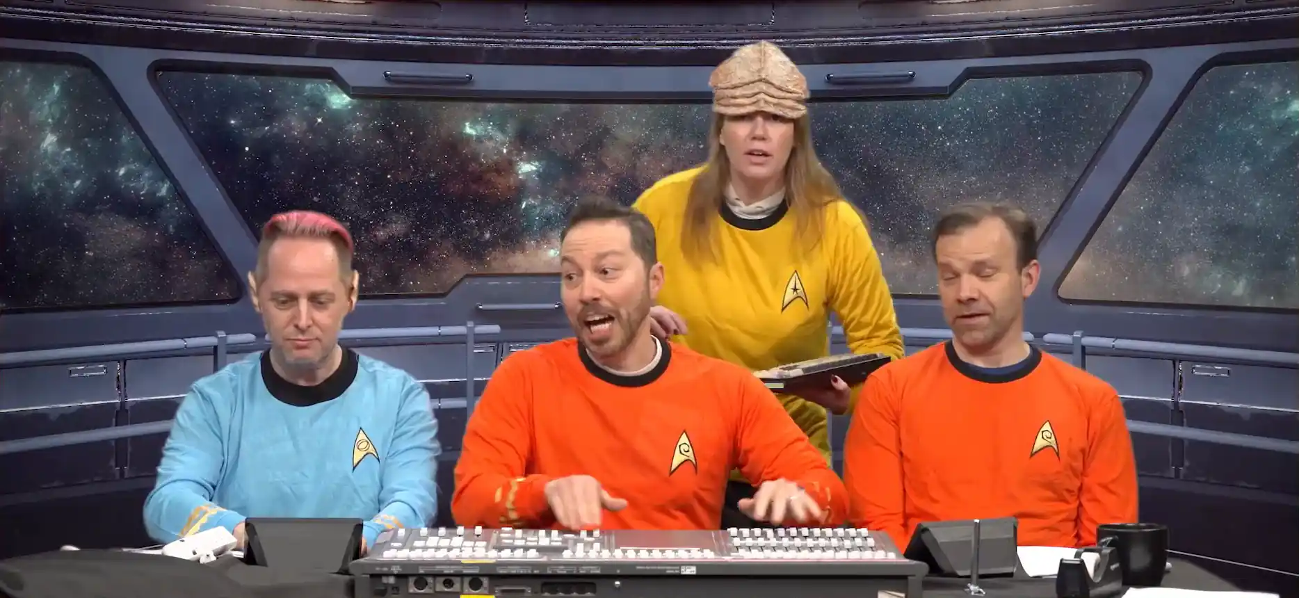 An animated image of Taliesin, Sam, Marisha, and Liam all dressed in Star Fleet uniforms on a green-screened star ship bridge. The camera is tilted to indicate the ship is shaking, and the cast is pretending to be thrown around. Taliesin wears a blue shirt and Spock ears. Sam and Liam wear red shirts. Marisha wears a yellow shirt and a thick leathery alien forehead prosthetic. Taliesin and Marisha look scared, while Sam and Liam laugh. A sound mixing board resembling a control panel sits in front of Sam.
